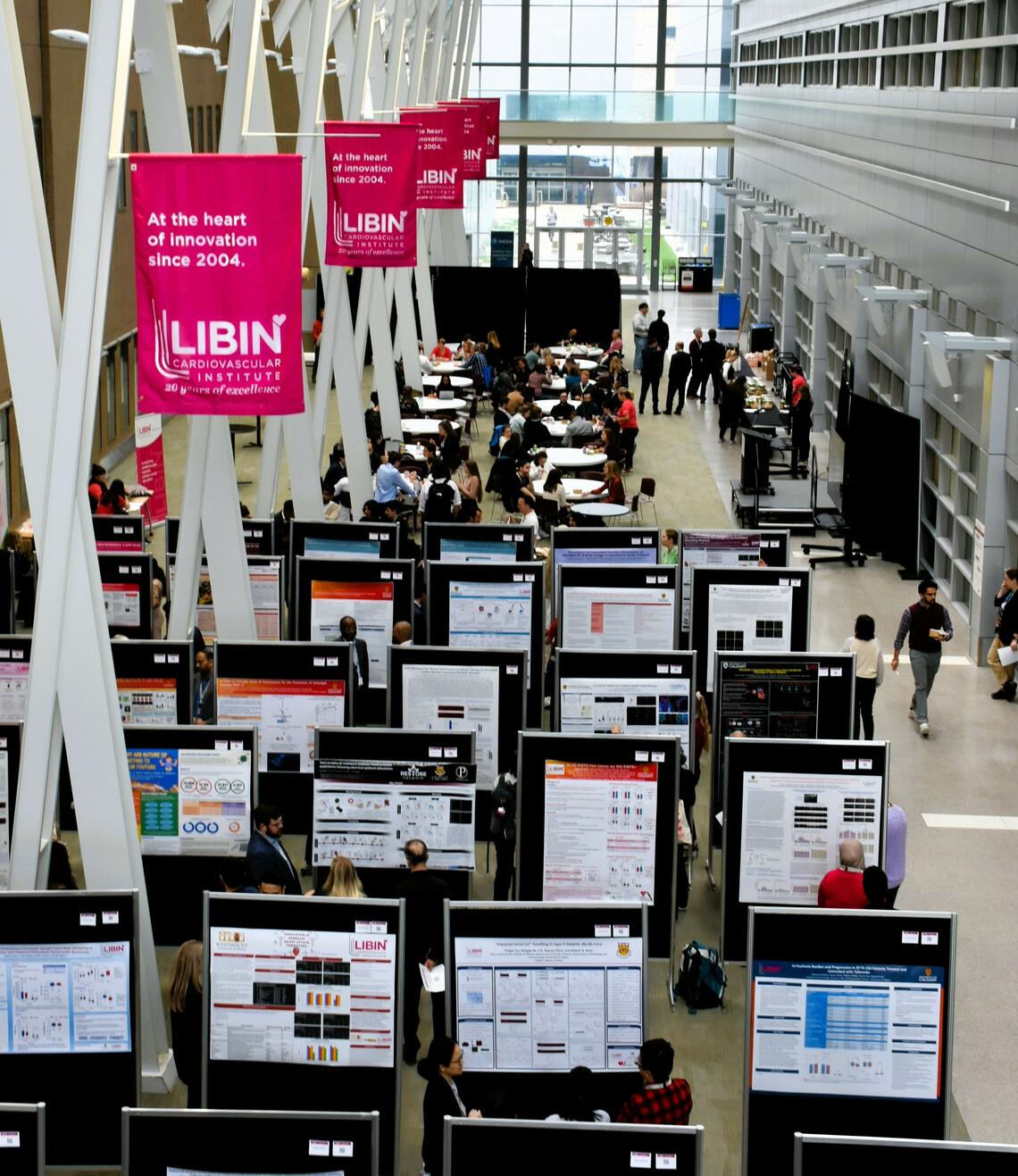 Black stands with research posters in a light filled room. Pink Libin Cardiovascular Institute flags above. 