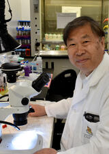 A man wearing a white lab coats sits in front of a microscope in a lab